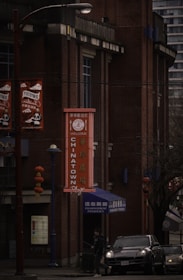 A street scene in Chinatown featuring a brick building with a vertical sign with the word 'Chinatown' in both English and Chinese characters. Red lanterns hang from a pole next to the building. A person is walking near a parked car under dim lighting, suggesting an overcast day.