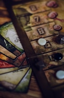 A close-up view of a board game setup, featuring a game board with numbered tiles and various game pieces, including dice and small tokens. Cards in different colors with intricate designs are fanned out beside the board.