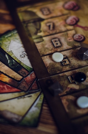 A close-up view of a board game setup, featuring a game board with numbered tiles and various game pieces, including dice and small tokens. Cards in different colors with intricate designs are fanned out beside the board.