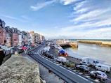 A coastal town features a row of colorful buildings along a winding road. The road runs parallel to the sea, where a harbor is visible with numerous boats docked. The sky is partly cloudy with hints of blue, and several cars are parked along the roadside. The area appears lively, with some people visiting market stalls under colorful canopies.