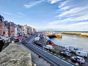 A coastal town features a row of colorful buildings along a winding road. The road runs parallel to the sea, where a harbor is visible with numerous boats docked. The sky is partly cloudy with hints of blue, and several cars are parked along the roadside. The area appears lively, with some people visiting market stalls under colorful canopies.