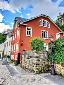 A picturesque street features a vibrant red house with white trim and a steep, dark roof. The house is set against a backdrop of lush greenery and a blue, cloud-streaked sky. A cobblestone path runs alongside the home, bordered by a low stone wall and lampposts. Potted plants add a touch of nature to the scene.