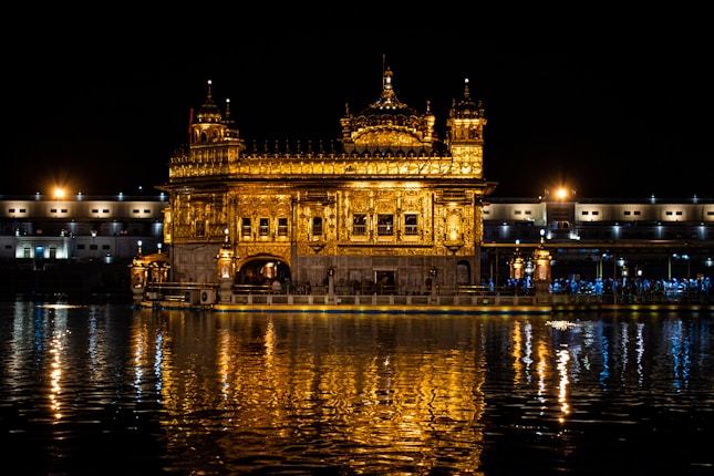 A magnificent golden temple illuminated at night, reflecting beautifully in the water below. It is surrounded by a calm, dark night sky, with subtle lighting along the perimeter of the temple complex.
