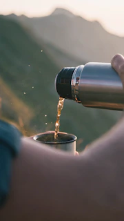 A person is pouring liquid from a metal thermos into a cup with a natural outdoor setting in the background. Sunlight creates a warm glow, highlighting the liquid being poured and emphasizing the serene setting.