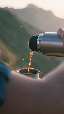 A person is pouring liquid from a metal thermos into a cup with a natural outdoor setting in the background. Sunlight creates a warm glow, highlighting the liquid being poured and emphasizing the serene setting.