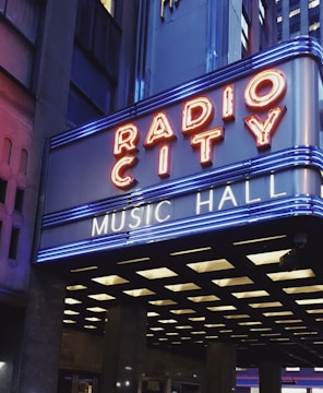 A brightly lit marquee displaying the words 'Radio City Music Hall' in neon lights, set against a backdrop of urban architecture. The marquee features red and blue neon lights, enhancing its visibility and vibrancy in the surrounding cityscape.