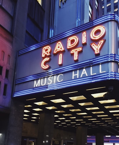 A brightly lit marquee displaying the words 'Radio City Music Hall' in neon lights, set against a backdrop of urban architecture. The marquee features red and blue neon lights, enhancing its visibility and vibrancy in the surrounding cityscape.