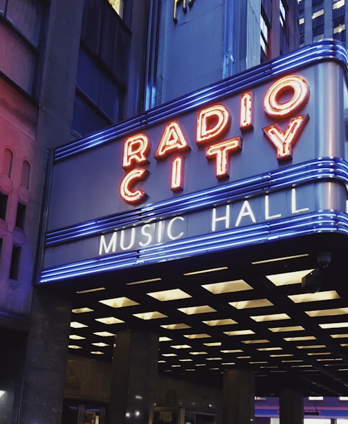 A brightly lit marquee displaying the words 'Radio City Music Hall' in neon lights, set against a backdrop of urban architecture. The marquee features red and blue neon lights, enhancing its visibility and vibrancy in the surrounding cityscape.