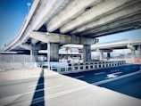 A concrete overpass with multiple lanes is visible, supported by large rectangular columns. The road below is marked with white arrows and bordered by metal railings. There is a chain-link fence and some signage in the background, along with urban elements.