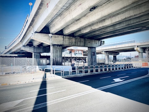 A concrete overpass with multiple lanes is visible, supported by large rectangular columns. The road below is marked with white arrows and bordered by metal railings. There is a chain-link fence and some signage in the background, along with urban elements.