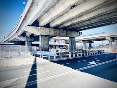 A concrete overpass with multiple lanes is visible, supported by large rectangular columns. The road below is marked with white arrows and bordered by metal railings. There is a chain-link fence and some signage in the background, along with urban elements.