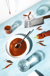 A captivating culinary scene featuring a vibrant setup on a light blue surface. At the center, a white plate holds a glossy brown sauce with a spoon resting on it. Nearby, a large red chili pepper and several smaller peppers are scattered, adding a touch of brightness. A glass jar filled with more of the brown sauce sits to one side, while an empty glass and a cleaver with a wooden handle lie nearby. A crystal glass contains more peppers, creating dynamic shadows across the surface.