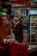 A person wearing a face mask stands inside a shop crowded with various packaged goods. Shelves filled with colorful products are visible, including beverages and snacks. A cooler on the right side contains drinks, and there is a counter with an electronic scale. The lighting is warm and cozy.