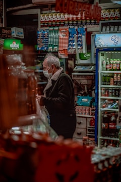 A person wearing a face mask stands inside a shop crowded with various packaged goods. Shelves filled with colorful products are visible, including beverages and snacks. A cooler on the right side contains drinks, and there is a counter with an electronic scale. The lighting is warm and cozy.