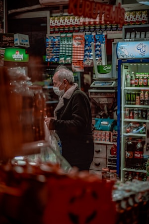 A person wearing a face mask stands inside a shop crowded with various packaged goods. Shelves filled with colorful products are visible, including beverages and snacks. A cooler on the right side contains drinks, and there is a counter with an electronic scale. The lighting is warm and cozy.