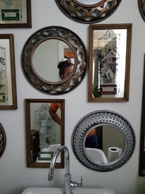 A wall decorated with various ornate mirrors and framed artworks featuring intricate designs. Some mirrors have detailed patterns around their edges. A sink with a modern faucet is positioned below the mirrors, and a roll of toilet paper is visible in the background. Part of a person is visible in two mirrors, including a part of their head and torso.