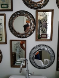 A wall decorated with various ornate mirrors and framed artworks featuring intricate designs. Some mirrors have detailed patterns around their edges. A sink with a modern faucet is positioned below the mirrors, and a roll of toilet paper is visible in the background. Part of a person is visible in two mirrors, including a part of their head and torso.