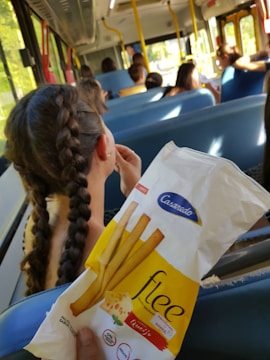 A group of people is seated on a bus with blue seats. The main focus is a hand holding a snack packet labeled 'Casarecdo, flee' with cheese-flavored breadsticks. In front, a person with braided hair is visible, looking toward the front of the bus.