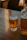 A glass of light beer with foam at the top, labeled with 'Upward Brewing Company', sits on a wooden table. To the right of the glass, there is a transparent pitcher with a similar light beer. The background includes softly lit furniture, creating a cozy atmosphere.
