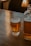A glass of light beer with foam at the top, labeled with 'Upward Brewing Company', sits on a wooden table. To the right of the glass, there is a transparent pitcher with a similar light beer. The background includes softly lit furniture, creating a cozy atmosphere.
