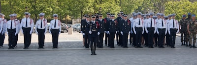 A large group of people in uniform is standing in neat rows and columns. The individuals appear to be in a formal formation, possibly a military or police parade. The uniforms are predominantly white shirts with epaulettes, dark pants, and hats. There is one person standing slightly ahead of the others, possibly in a leadership position. The background includes green trees and parked cars.
