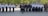 A large group of people in uniform is standing in neat rows and columns. The individuals appear to be in a formal formation, possibly a military or police parade. The uniforms are predominantly white shirts with epaulettes, dark pants, and hats. There is one person standing slightly ahead of the others, possibly in a leadership position. The background includes green trees and parked cars.