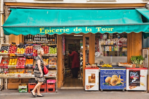 A small French grocery store with an awning displaying the name 'Epicerie de la Tour'. The front of the store is lined with crates of fresh fruits such as apples and citrus. A variety of products are stacked inside, visible through the glass door. A woman with gray hair carrying a red bag and wearing a black and white dress walks by. Posters and advertisements cover the freezer at the entrance.
