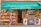 A small French grocery store with an awning displaying the name 'Epicerie de la Tour'. The front of the store is lined with crates of fresh fruits such as apples and citrus. A variety of products are stacked inside, visible through the glass door. A woman with gray hair carrying a red bag and wearing a black and white dress walks by. Posters and advertisements cover the freezer at the entrance.