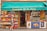 A small French grocery store with an awning displaying the name 'Epicerie de la Tour'. The front of the store is lined with crates of fresh fruits such as apples and citrus. A variety of products are stacked inside, visible through the glass door. A woman with gray hair carrying a red bag and wearing a black and white dress walks by. Posters and advertisements cover the freezer at the entrance.
