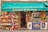 A small French grocery store with an awning displaying the name 'Epicerie de la Tour'. The front of the store is lined with crates of fresh fruits such as apples and citrus. A variety of products are stacked inside, visible through the glass door. A woman with gray hair carrying a red bag and wearing a black and white dress walks by. Posters and advertisements cover the freezer at the entrance.