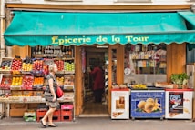 A small French grocery store with an awning displaying the name 'Epicerie de la Tour'. The front of the store is lined with crates of fresh fruits such as apples and citrus. A variety of products are stacked inside, visible through the glass door. A woman with gray hair carrying a red bag and wearing a black and white dress walks by. Posters and advertisements cover the freezer at the entrance.