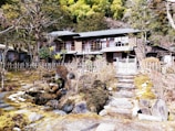 A traditional wooden house is set in a serene garden surrounded by a rustic wooden fence. The garden has stone pathways and steps leading up to the house, with lush greenery and trees in the background. The house features a classic architectural style with large windows and wooden sliding doors.