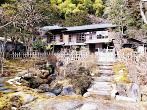 A traditional wooden house is set in a serene garden surrounded by a rustic wooden fence. The garden has stone pathways and steps leading up to the house, with lush greenery and trees in the background. The house features a classic architectural style with large windows and wooden sliding doors.