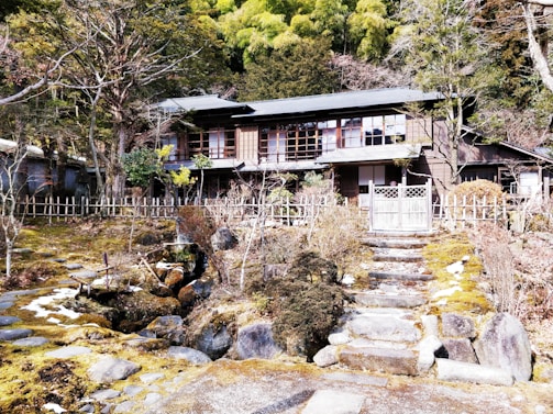 A traditional wooden house is set in a serene garden surrounded by a rustic wooden fence. The garden has stone pathways and steps leading up to the house, with lush greenery and trees in the background. The house features a classic architectural style with large windows and wooden sliding doors.