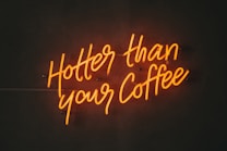 Neon sign with script lettering reading 'Hotter than your Coffee' glowing in a dark environment.