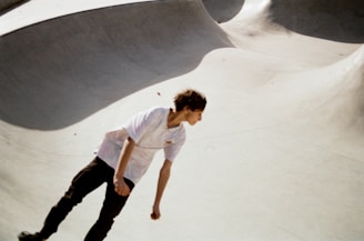 A young person is skateboarding in a concrete skate park, which features smooth, curved surfaces. The lighting suggests it is a sunny day, casting distinct shadows across the park. The skateboarder is wearing a white t-shirt and dark pants.