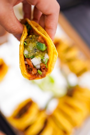 A hand holds a taco filled with ingredients like diced onions, cilantro, and a green salsa. The taco shell is yellow and crispy, and the filling appears to contain seasoned meat.