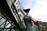 A sign with large letters spelling 'ROCK SHOP' is prominently featured on the facade of a modern building. The building has a lot of glass panels and metal elements with an industrial aesthetic. The logo of a well-known restaurant or cafe is also visible, suggesting a themed retail or dining establishment.