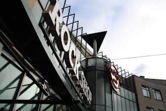 A sign with large letters spelling 'ROCK SHOP' is prominently featured on the facade of a modern building. The building has a lot of glass panels and metal elements with an industrial aesthetic. The logo of a well-known restaurant or cafe is also visible, suggesting a themed retail or dining establishment.