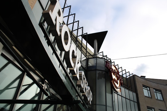 A sign with large letters spelling 'ROCK SHOP' is prominently featured on the facade of a modern building. The building has a lot of glass panels and metal elements with an industrial aesthetic. The logo of a well-known restaurant or cafe is also visible, suggesting a themed retail or dining establishment.