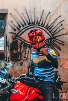 A person wearing a vibrant red and orange helmet with goggles sits on a red motorcycle. They are in front of a wall displaying a large graffiti of an eye. The individual is gesturing with their hand, wearing a striped shirt in blue and yellow hues.