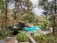 A swimming pool surrounded by lush greenery and trees, featuring a thatched-roof structure by the poolside. The area is peaceful and serene, suggesting a tropical or resort-like setting.