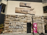 A restaurant facade featuring rustic wooden paneling and a bold sign with the name 'good spoon'. The establishment offers all-day brunch and dinner, specializing in grill and wine. Above the door is another sign for 'Atelier Mokuba Osaka Gallery' on a wooden backdrop. A smaller, vibrant pink poster with various images is displayed to the right of the entrance.