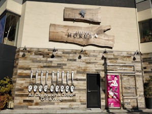 A restaurant facade featuring rustic wooden paneling and a bold sign with the name 'good spoon'. The establishment offers all-day brunch and dinner, specializing in grill and wine. Above the door is another sign for 'Atelier Mokuba Osaka Gallery' on a wooden backdrop. A smaller, vibrant pink poster with various images is displayed to the right of the entrance.