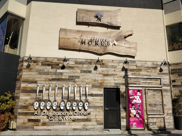 A restaurant facade featuring rustic wooden paneling and a bold sign with the name 'good spoon'. The establishment offers all-day brunch and dinner, specializing in grill and wine. Above the door is another sign for 'Atelier Mokuba Osaka Gallery' on a wooden backdrop. A smaller, vibrant pink poster with various images is displayed to the right of the entrance.