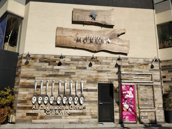 A restaurant facade featuring rustic wooden paneling and a bold sign with the name 'good spoon'. The establishment offers all-day brunch and dinner, specializing in grill and wine. Above the door is another sign for 'Atelier Mokuba Osaka Gallery' on a wooden backdrop. A smaller, vibrant pink poster with various images is displayed to the right of the entrance.