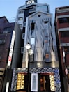 A modern hotel building with a unique facade featuring wave-like patterns. The exterior combines metal and glass elements with illuminated signs and decorative circular motifs near the entrance. The architecture stands out with its contemporary design and lighting, set in an urban environment.