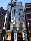 A modern hotel building with a unique facade featuring wave-like patterns. The exterior combines metal and glass elements with illuminated signs and decorative circular motifs near the entrance. The architecture stands out with its contemporary design and lighting, set in an urban environment.