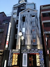 A modern hotel building with a unique facade featuring wave-like patterns. The exterior combines metal and glass elements with illuminated signs and decorative circular motifs near the entrance. The architecture stands out with its contemporary design and lighting, set in an urban environment.
