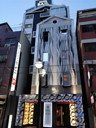 A modern hotel building with a unique facade featuring wave-like patterns. The exterior combines metal and glass elements with illuminated signs and decorative circular motifs near the entrance. The architecture stands out with its contemporary design and lighting, set in an urban environment.